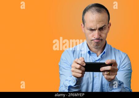 Mature man in formal wear looking at his smartphone and biting his upper lip. Orange background. Stock Photo