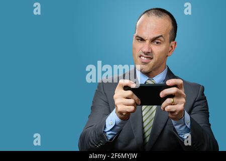 Mature man with suit and tie, playing on his smartphone and making a funny angry face. Blue background. Stock Photo