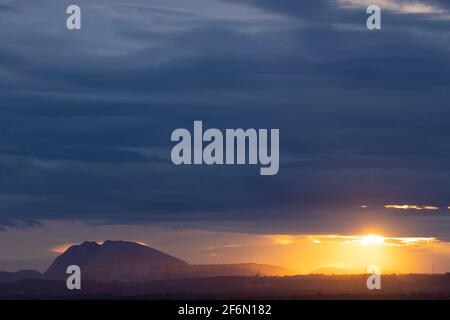 A Panoramic landscape view of dark clouds on the horizon and a hill in the bottom and the golden  sun rays passing through them Stock Photo