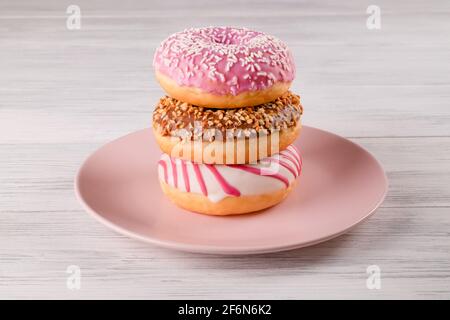 Three sugar-coated donuts are stacked on a pink ceramic plate on a light-colored wooden surface. Selective focus. Stock Photo