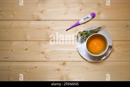 A cup of herbal tea made of thyme. With the movement of infusion waves of divorces. With a floral bouquet on a saucer. The thermometer has 37.7 on display. On a countertop light wooden boards. Stock Photo