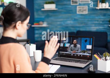 Student waving hand to her university teacher during videocall meeting. Caucasian female discussing about remote education because of coronavirus quarantine while sitting at desk in living room Stock Photo