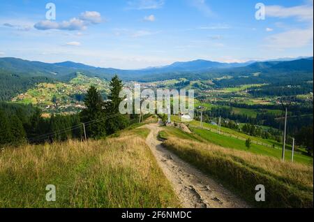 Dirt road in a village leads to the town in mountains. Countryside road and wide view of small town in a valley and mountains around. Vorokhta Stock Photo