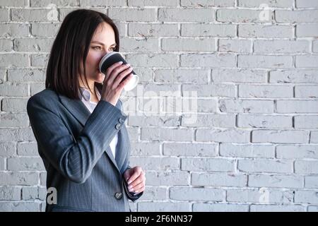 Smiling company manager in white blouse and grey classic costume drinks coffee from paper cup standing against brick wall before work copyspace Stock Photo