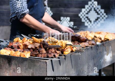 Tasty fried pieces of meat on skewers are roasted on grill at festival outdoor. Chef in black apron prepares barbecue. Shish kebab national cuisine of Stock Photo