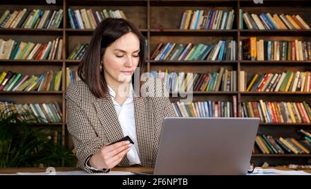 Online shopper in brown jacket enters credit card number typing on grey laptop sitting at table against large racks with books at home Stock Photo