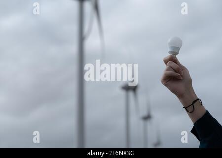 Hand portrait of caucasian woman holding an energy saving bulb, in background a turbines  . Renewable energy, future and electricity concept. Stock Photo