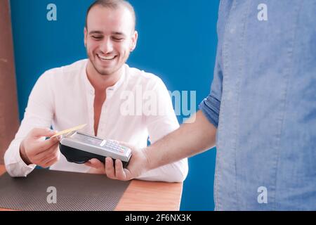 Young smiling man with card holding it over payment terminal while paying for her order in coffee restaurant. Technology concept. Stock Photo