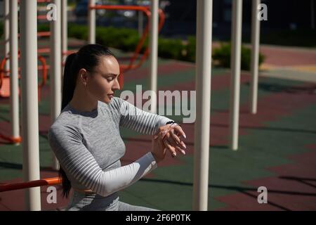 Young sportswoman checking her fitness tracker, pulse and heart rate, lost calories during outdoor workout, standing ona sports ground. Sports trainin Stock Photo