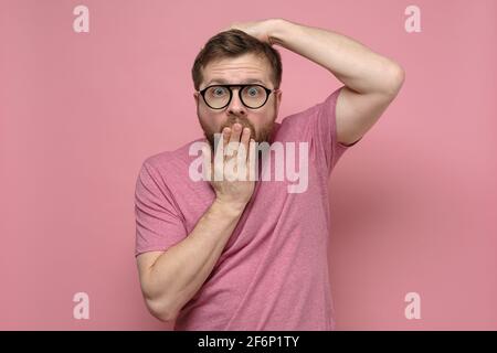 Shocked, strange man with glasses in fright covers mouth with one hand, put the other on head and looks with big eyes.  Stock Photo