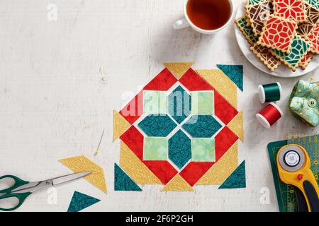 Pieces of fabric laid out in the shape of a patchwork block, a  heap of cookies with a pattern imitating a patchwork block, a cup of tea, sewing and q Stock Photo