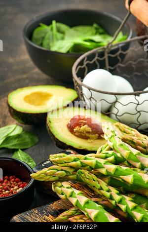 Bunch of fresh green organic asparagus spinach, avocado, chicken eggs and pepper seasoning on black old wooden background, top view. Food cooking back Stock Photo
