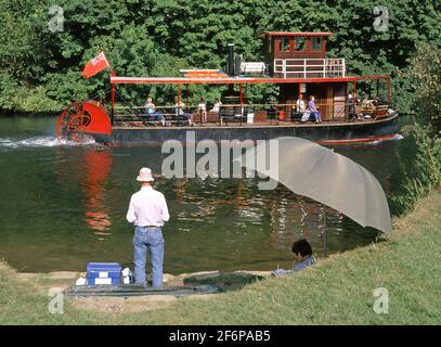 People on Thames paddle steamer tour boat Lucy Fisher passes man fishing on river bank with companion relaxing in shade of umbrella Egham Surrey UK Stock Photo
