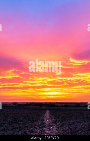 Dramatic dawn sky, orange yellow and mauve, over a landscape with, in the foreground, a field and muddy path running into the center of the picture. Stock Photo