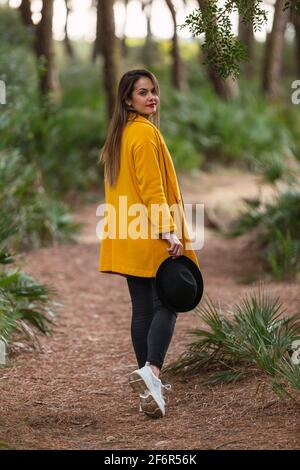 Back view of elegant young woman in yellow casual coat in the forest. Cute model walks in the park in spring against background of nature. Stock Photo