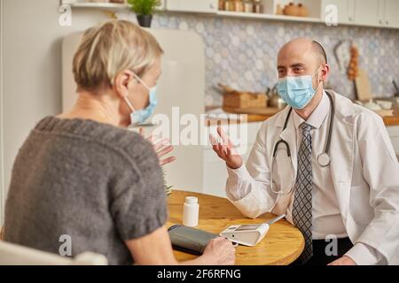A male doctor wearing a protective mask advises an elderly woman wearing a protective mask at her home. Stock Photo