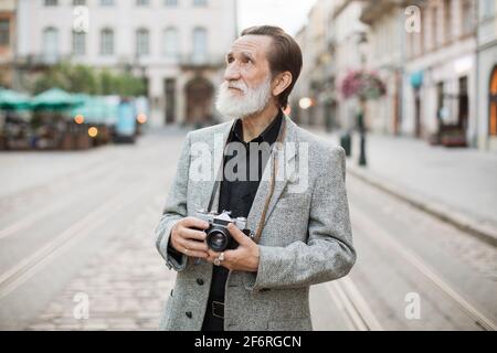 Senior bearded man standing on city street with retro camera on neck. Male tourist in stylish clothes enjoying beautiful architecture of old town. Stock Photo
