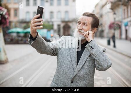 Happy mature man using modern smartphone for video chat on fresh air. Stylish bearded pensioner smiling and doing a selfie during online conversation. Urban space. Stock Photo