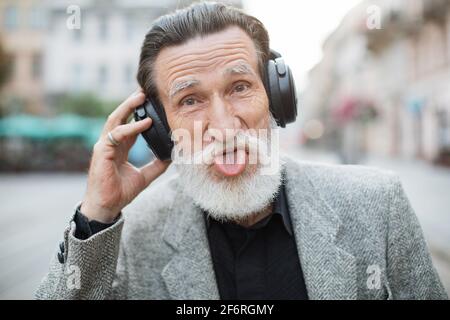 Funny elder man with grey beard wearing wireless headphones and showing tongue while standing on city street. Happy pensioner listening music outdoors. Stock Photo