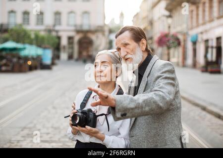 Beautiful mature woman with retro photo camera on neck listening her bearded husband that hugging her. Senior family enjoying travelling time during retirement. Spending time together. Stock Photo