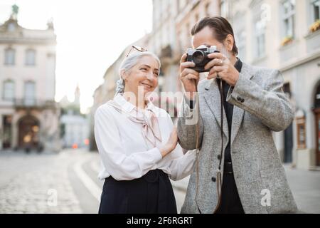 Lovely senior couple in elegant clothes walking on city street and taking pictures on retro camera. Happy mature tourists spending time together during holidays. Stock Photo
