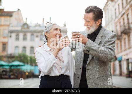 Side view of smiling aged couple in stylish outfits toasting with carton cups of hot coffee while standing on street. Happy mature family smiling and looking kindly on each other. Stock Photo