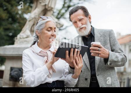 Bearded senior man holding digital tablet while his lovely wife pointing on screen. Two mature people sitting outdoors, drinking coffee and using modern gadget. Stock Photo