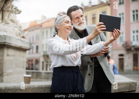 Smiling aged man and woman taking selfie on digital tablet while relaxing on fresh air. Senior couple holding cups of coffee. Family enjoying retirement. Stock Photo