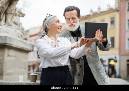 Bearded senior man holding digital tablet while his lovely wife pointing on screen. Two mature people sitting outdoors, drinking coffee and using modern gadget. Stock Photo