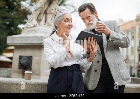Bearded senior man holding digital tablet while his lovely wife pointing on screen. Two mature people sitting outdoors, drinking coffee and using modern gadget. Stock Photo