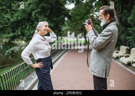 Smiling aged man using retro camera for taking photo of his charming lovely wife. Senior couple walking at green park and spending time together. Stock Photo