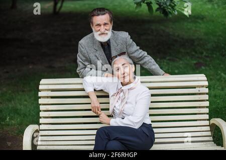 Attractive aged woman sitting on wooden bench while her bearded husband standing behind. Stylish mature couple posing at green park. Happy family. Stock Photo