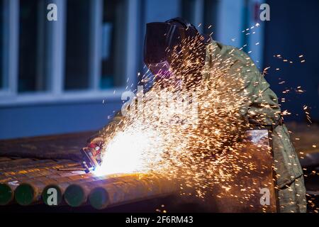 Welder worker in protective mask. Welding steel tubes on manufacturing plant. Sparks and smoke. On blue factory background. Stock Photo