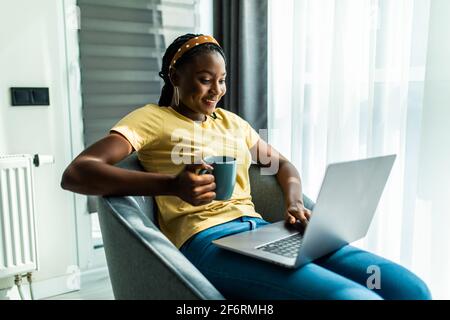 Happy Afro-American millennial woman sitting on chair, watching webinar, working online on laptop, talking in video chat, drinking tea or coffee. Stock Photo