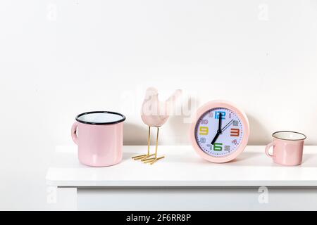 an alarm clock showing seven o'clock, cup, a pink bird figurine, a books on the table. Stock Photo