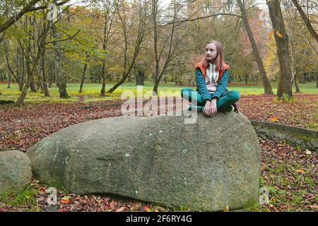 School age girl in turquoise and orange clothes sits on a huge shapeless boulder in an old autumn park, color editing photo Stock Photo