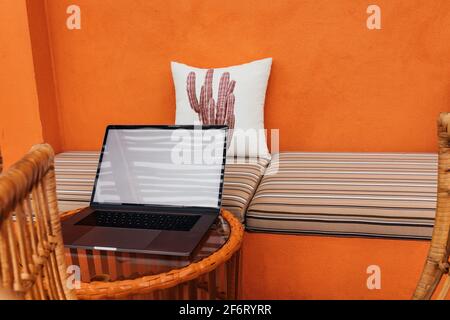A laptop on a glass table, a pillow against the background of an orange textured wall with a striped sofa in a modern cafe. Copy space. place to inser Stock Photo