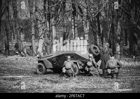 Group Of Reenactors Dressed As Russian Soviet Red Army Soldiers Of World War II Go On Offensive Under Cover Of Armored Soviet Scout Car. Historical Stock Photo