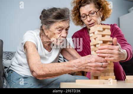 Retired mother and daughter spend time together at home, playing board game and caressing dachshund dog. Caucasian senior woman builds tower of wooden Stock Photo