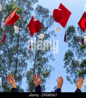 Hands of students throwing red graduation hats and masks into the air over a blue sky . Graduation and social distancing Stock Photo