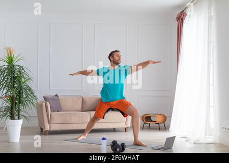 A middle aged man works out at home with an online tutorial. He performs a balance exercise in front of a laptop monitor. Stock Photo