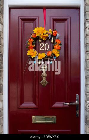 An orange and yellow floral Easter wreath on a red, paneled, wooden front door.  Door furniture (number, handle, letterbox) is in brass. Stock Photo