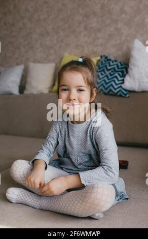a little girl in a gray dress sits on the sofa and smiles. home environment Stock Photo