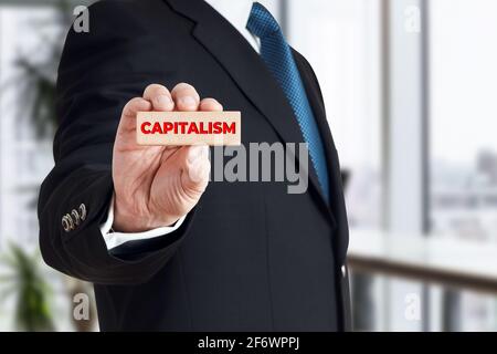 Businessman shows a wooden block with the word capitalism. Global corporate capitalism concept. Stock Photo