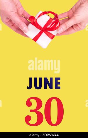 June 30th. Festive Vertical Calendar With Hands Holding White Gift Box With Red Ribbon And Calendar Date 30 June On Illuminating Yellow Background.Sum Stock Photo