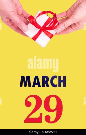 March 29th. Festive Vertical Calendar With Hands Holding White Gift Box With Red Ribbon And Calendar Date 29 March On Illuminating Yellow Background. Stock Photo