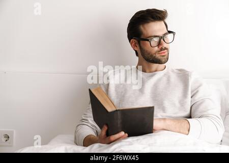 Focused young man in eyeglasses reading book while resting in bed at home Stock Photo