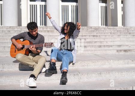 A young latin couple playing the guitar and singing sitting on stairs. Millennial generation. Stock Photo