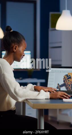 African engineer working late at night on a 3D model of industrial gears at laptop doing overtime in start up office. Freelancer studying prototype idea on pc showing cad software on device display Stock Photo