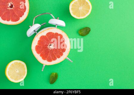 Half a grapefruit on the dial of the alarm clock and sliced fresh fruits on the green surface Stock Photo
